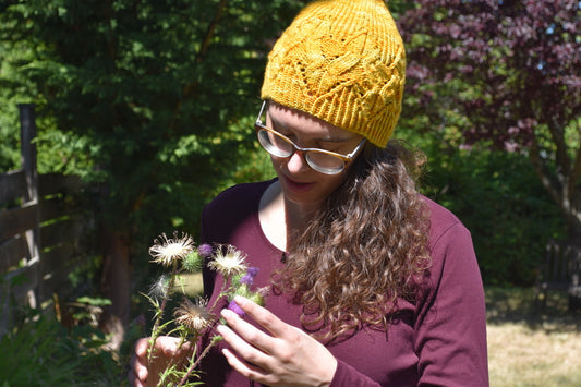 Woman with curly hair and glasses wearing a yellow knit hat and maroon shirt holding wildflowers outdoors