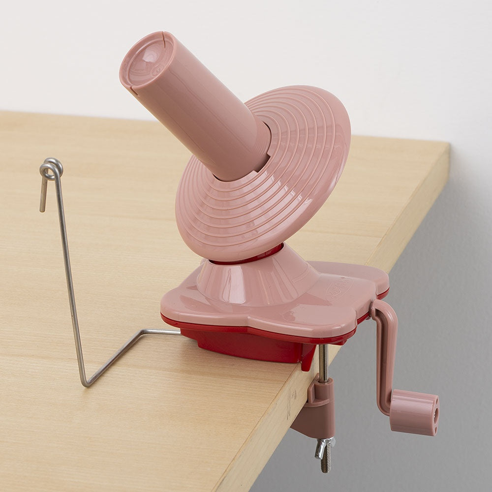 Pink plastic yarn ball winder clamped to light wooden desk with metal guide and hand crank