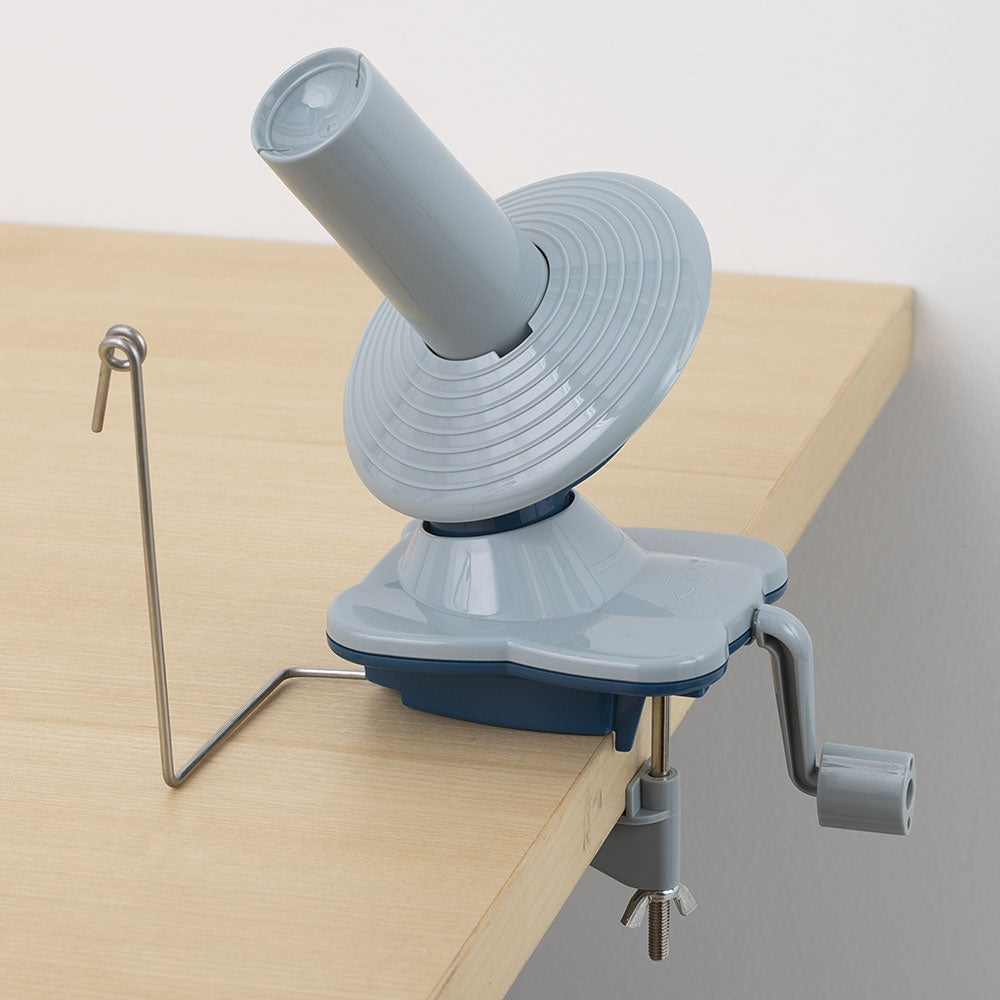 Gray handheld rotary yarn ball winder clamped to light wooden table edge with metal yarn guide