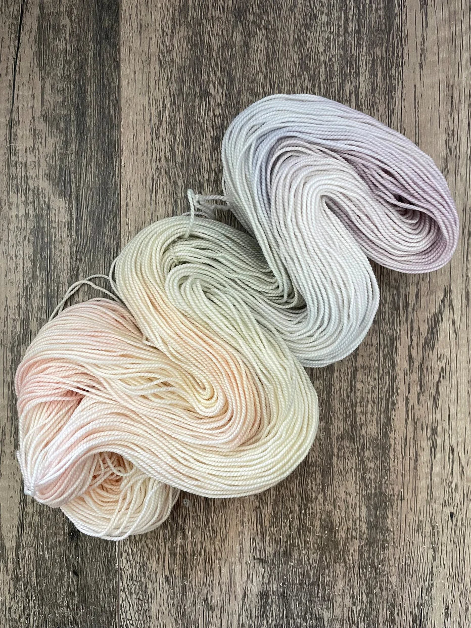 Two skeins of pastel hand-dyed yarn in soft peach, green, white, and lavender on wooden background
