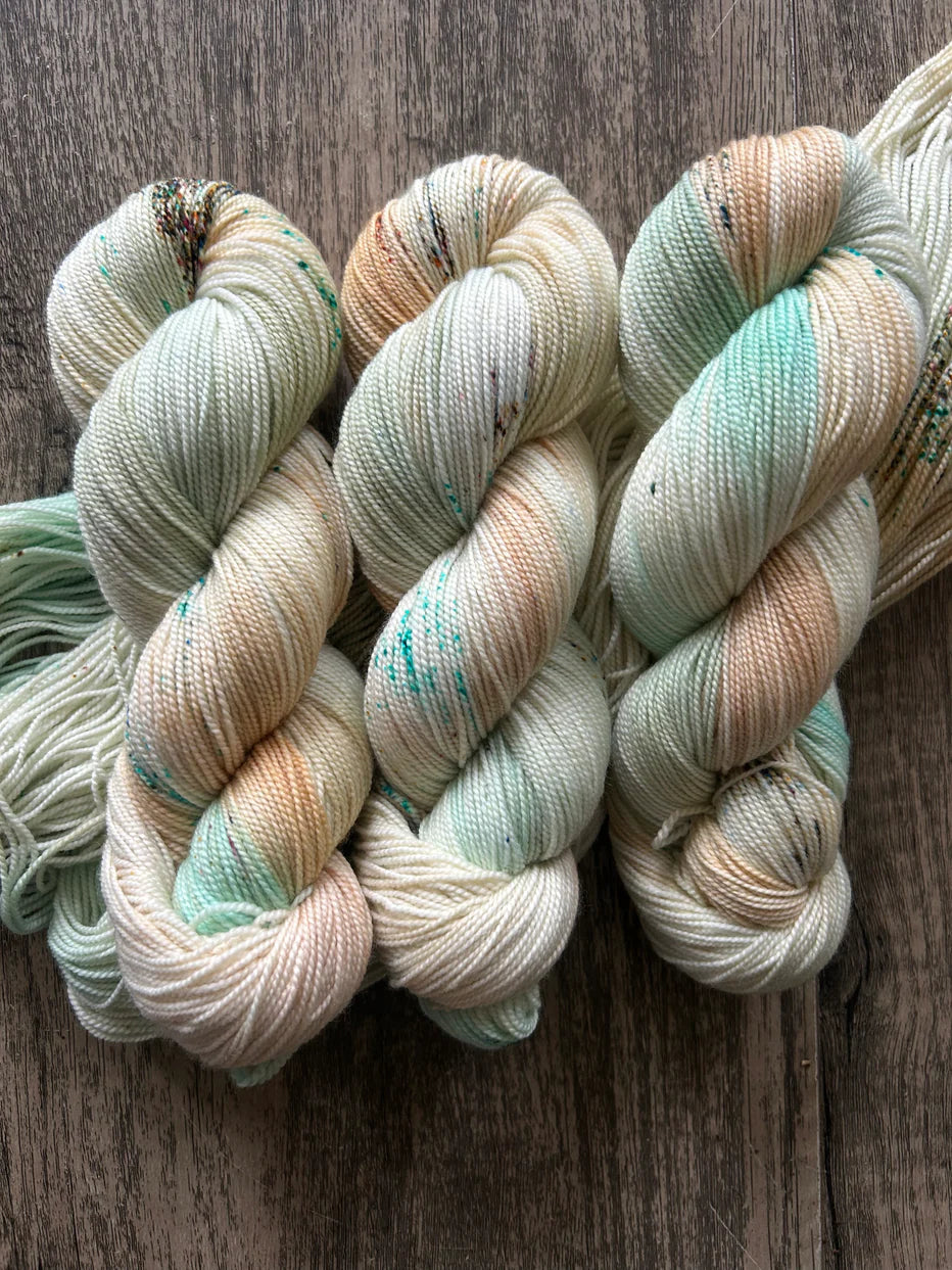 Three skeins of hand-dyed yarn in soft cream, mint green, and beige tones on rustic wooden surface