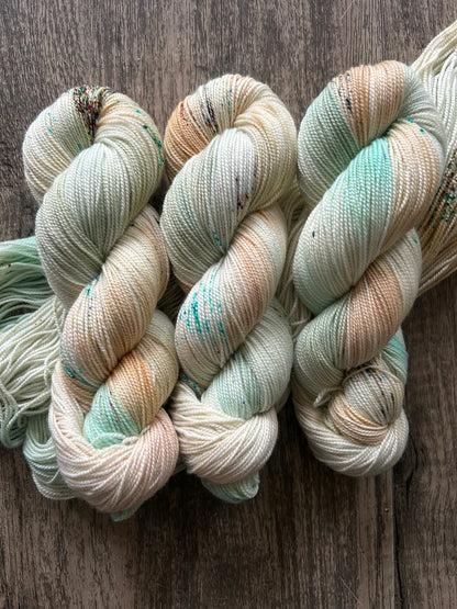 Three skeins of hand-dyed yarn in soft cream, mint green, and beige tones on rustic wooden surface